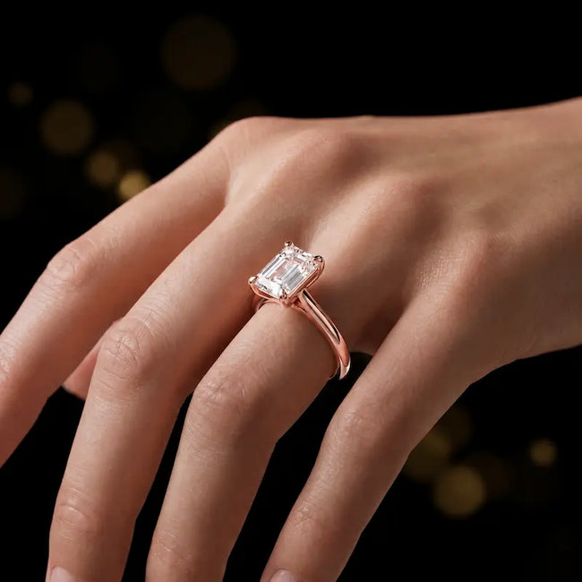 1.5 carat emerald cut diamond solitaire engagement ring in an 18kt rose gold 4 claw setting worn on a hand, by TJ Diamond