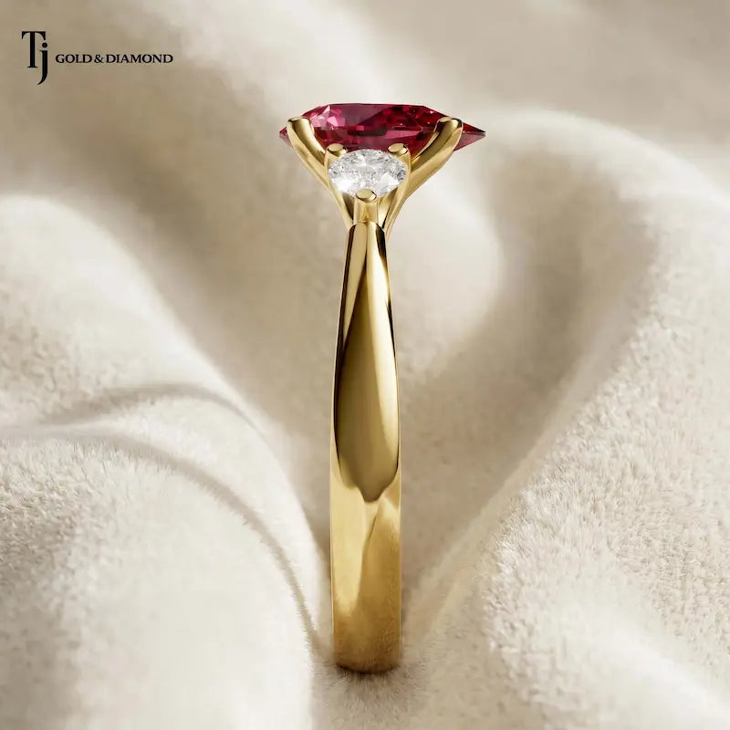 Side profile of a pear cut lab-created ruby trilogy engagement ring in 18kt yellow gold with pear cut lab-grown diamond side stones and a four claw basket setting.