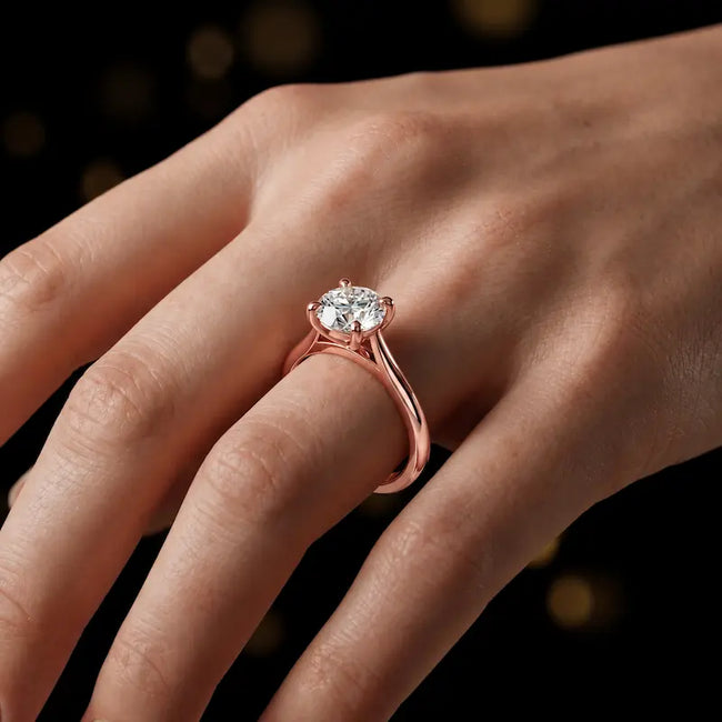 Round brilliant cut diamond solitaire engagement ring in an 18kt rose gold 4 claw setting worn on a hand, by TJ Diamond