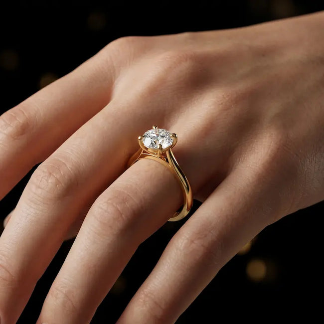 Round brilliant cut diamond solitaire engagement ring in an 18kt yellow gold 4 claw setting worn on a hand, by TJ Diamond