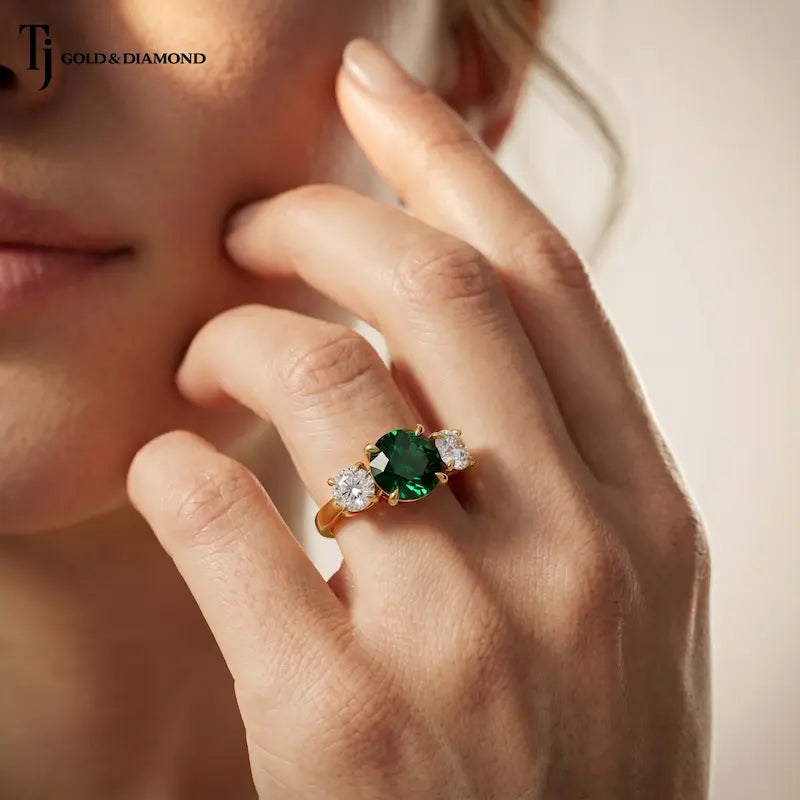 Round cut lab-created emerald trilogy engagement ring in 18kt yellow gold with round brilliant lab-grown diamond side stones, worn on hand.
